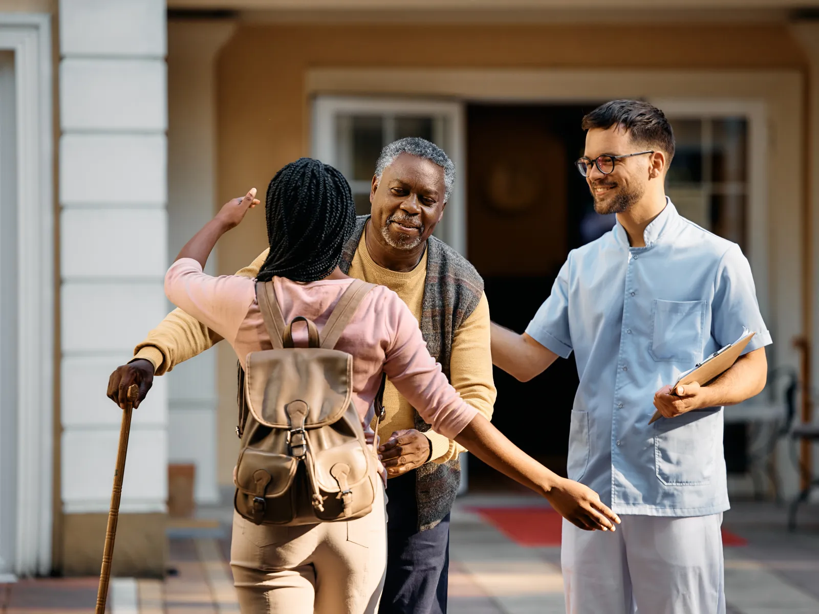 A caregiver welcomes an elderly man with a cane while another staff member smiles in a warm setting.